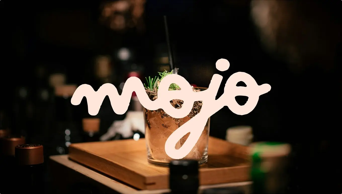 Le lettrage Mojo en néon beige devant un cocktail posé sur un plateau en bois, dans un bar.
